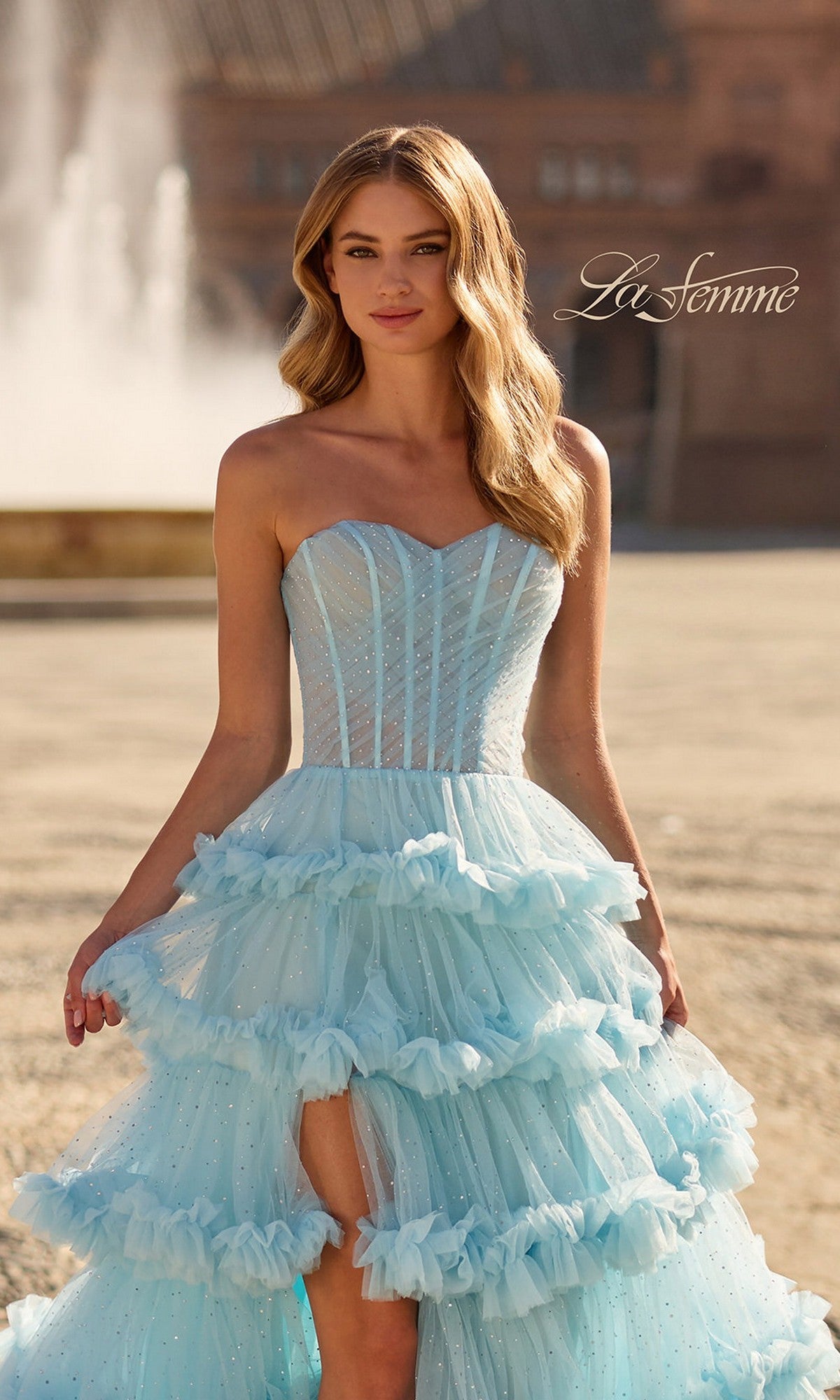 A woman stands outdoors near a fountain wearing the La Femme 33812 long prom dress, featuring a strapless corset bodice, ruffled tulle, and a high-low hem.